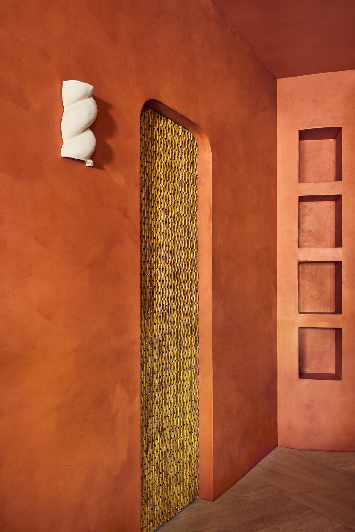 Murs à la chaux terracotta et rideaux en perles de bois, appliques céramique Babka Aurélie di Fretti. Comptoir en pierre avec étagères encastrées en bois et appliques céramique Babka d'Aurélie Fretti. Projet Love Café à Paris réalisé par Batiik Studio.