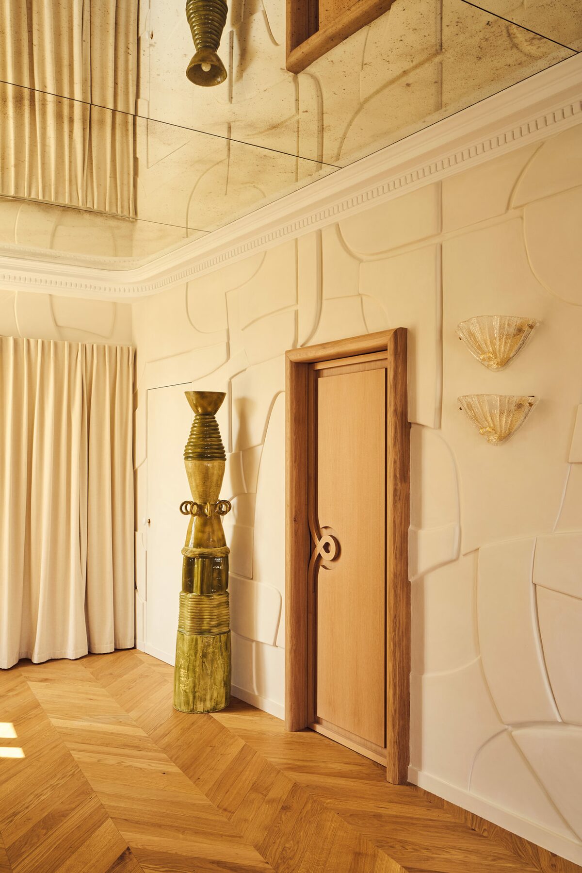 Entrée dans appartement haussamannien avec miroir au plafond, parquet chevrons, applique en verre, rideau blanc crème pour cacher rangement. Projet Joséphine à Paris signé Batiik Studio.