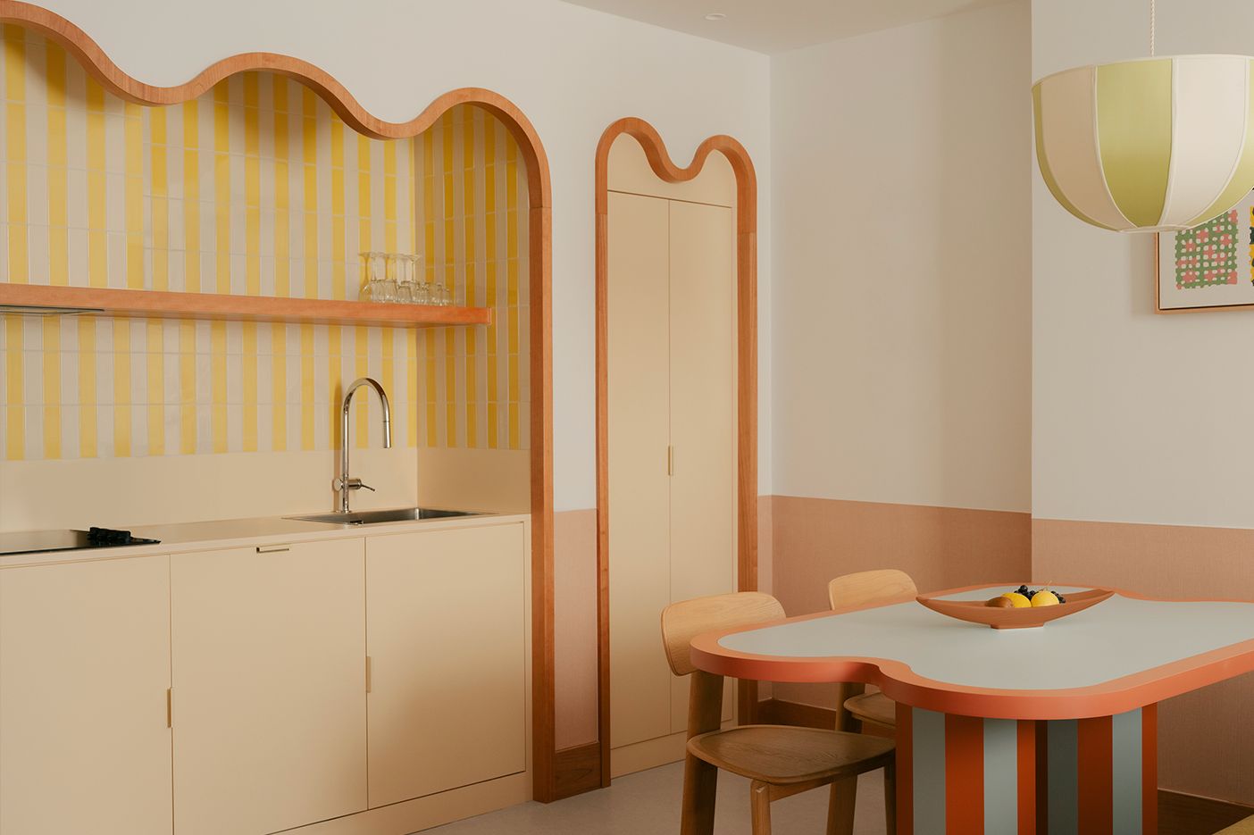 Salle à manger avec carreaux de faïence rayures blancs et jaunes. Encadrement en bois courbé. Style doux et méditerranéen, fantaisiste. Table de salle à manger avec pied rayé bleu clair et orange. Suspension ovale tissu rayé jaune vert et blanc. Projet hôtelier Croisette d'Edgar Suite à Cannes réalisé par Batiik Studio.