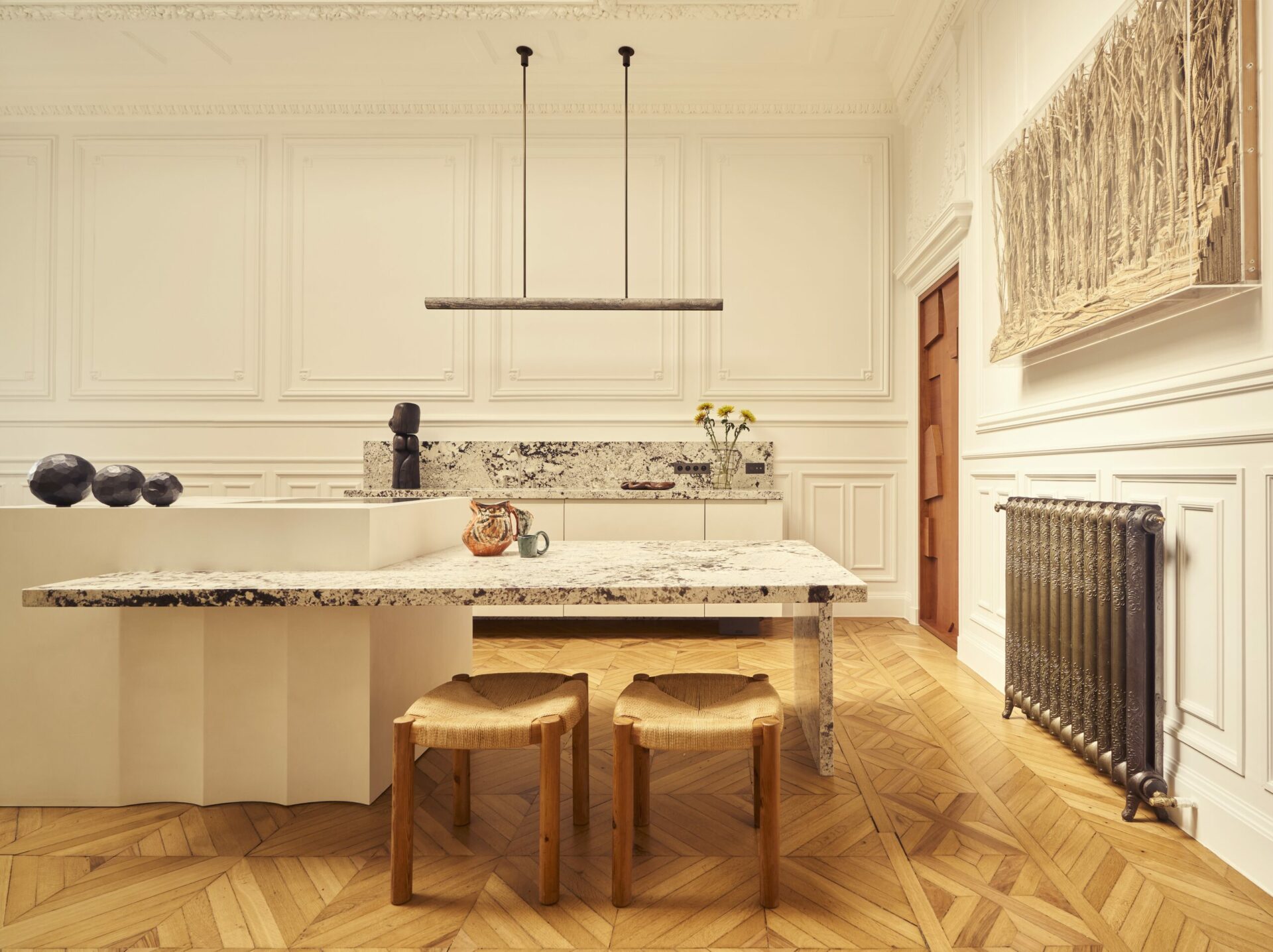 Cuisine en corian et granit avec plan de travail mural et îlot central éclairé par une verrière d’époque. Chaises Kelly de Frederic Pellenq vendues par la galerie Kolkhoze. Table de salle à manger réalisée sur-mesure par Batiik Studio. Projet Gabriel réalisé par Batiik Studio.