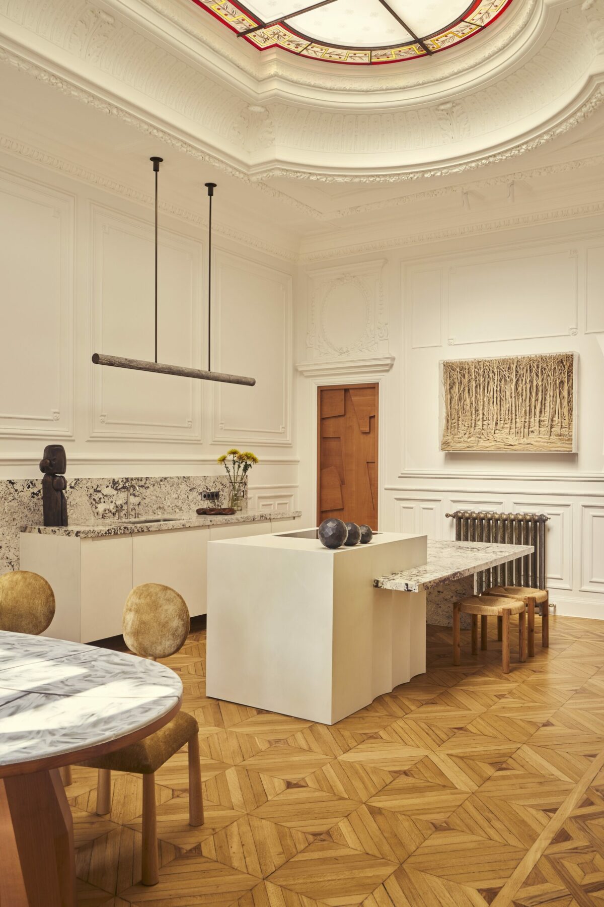 Cuisine en corian et granit avec plan de travail mural et îlot central éclairé par une verrière d’époque. Chaises Kelly de Frederic Pellenq vendues par la galerie Kolkhoze. Table de salle à manger réalisée sur-mesure par Batiik Studio. Projet Gabriel réalisé par Batiik Studio.