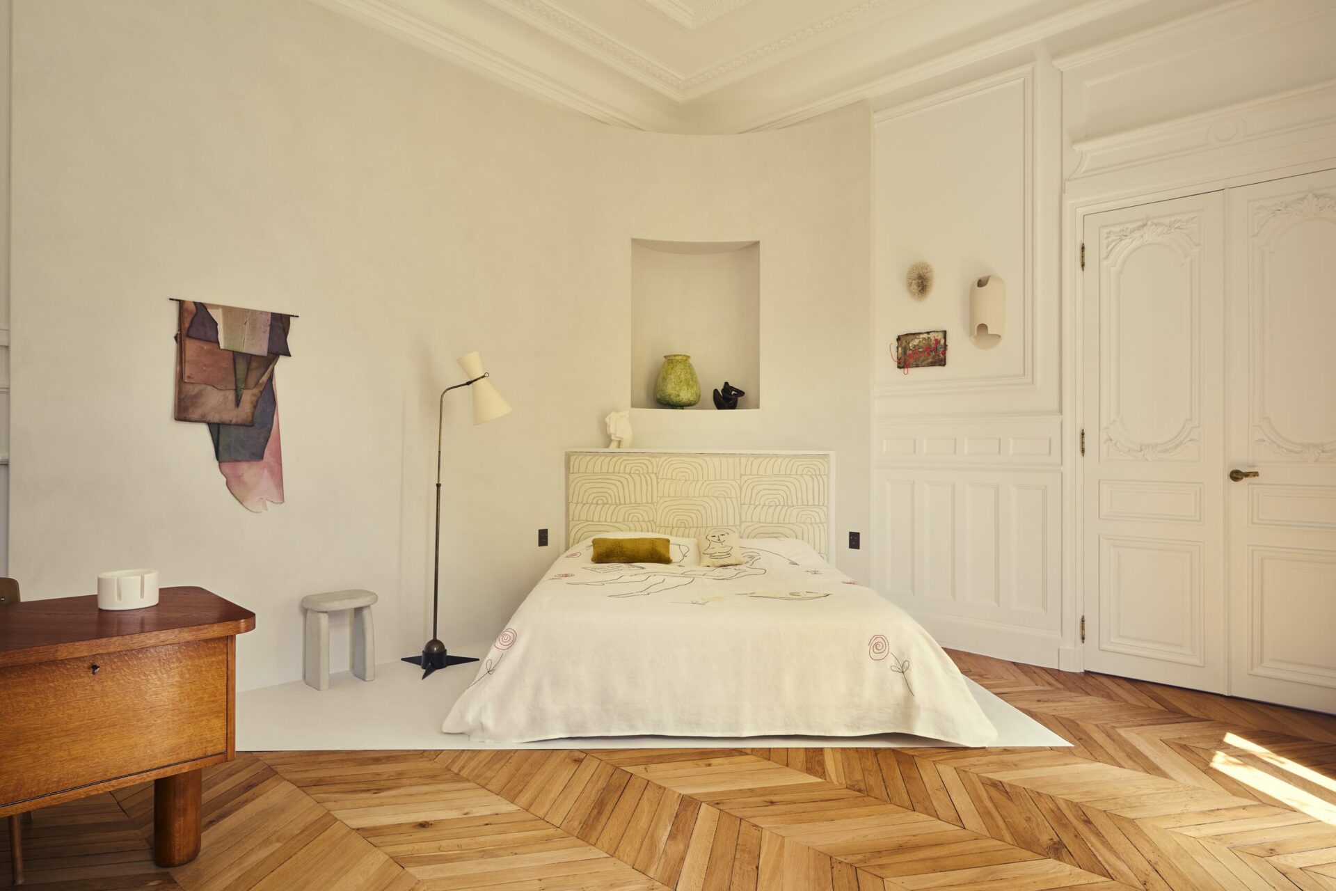 Chambre parentale installée en biais est délimitée par une résine blanche au sol. Une cloison qui s’enroule derrière la tête de lit afin de combler l’angle et créer une niche où exposer des éléments décoratifs. Projet Gabriel réalisé par Batiik Studio. Dessus de lit brodé de Sarah Espeute d'oeuvres sensibles, tabouret Window of Bo Bardi de Linde freya tangelder. Tissu tête de lit Schumacher, applique Johanna de Clisson, vase vert Camille Romagnani, oeuvres textiles Natalia Jaime-Cortez.